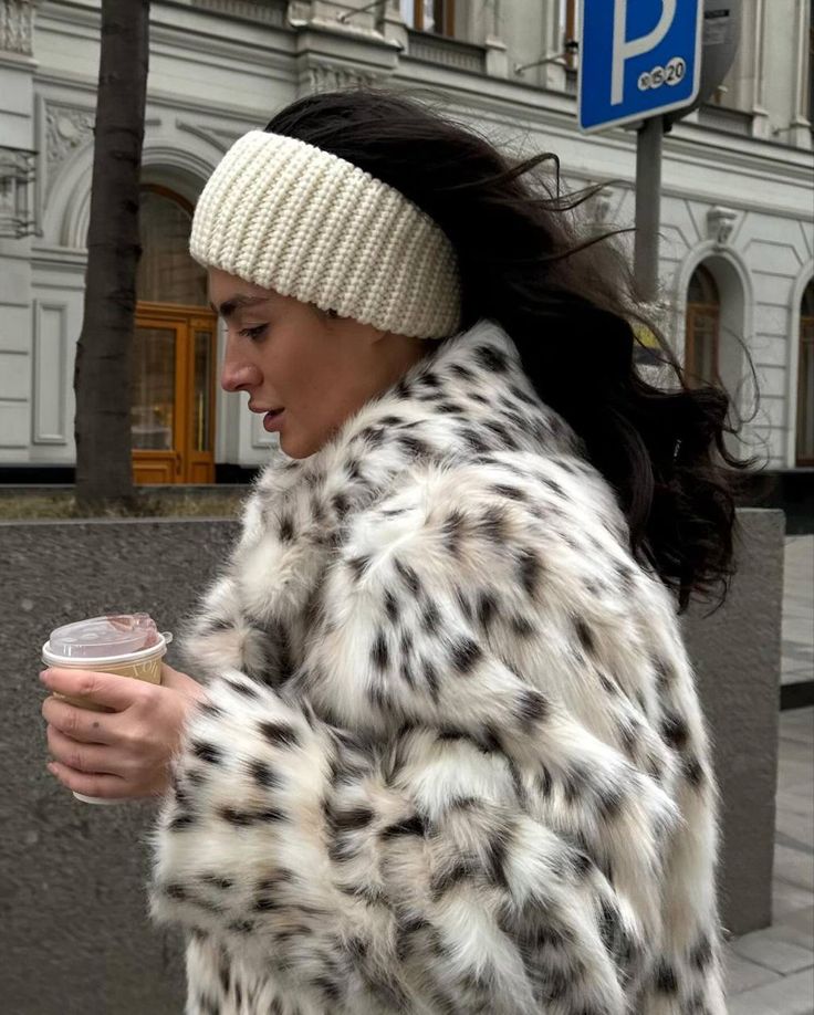 Woman in a spotted fur coat, wearing a knitted headband, holding a coffee cup on a city street.