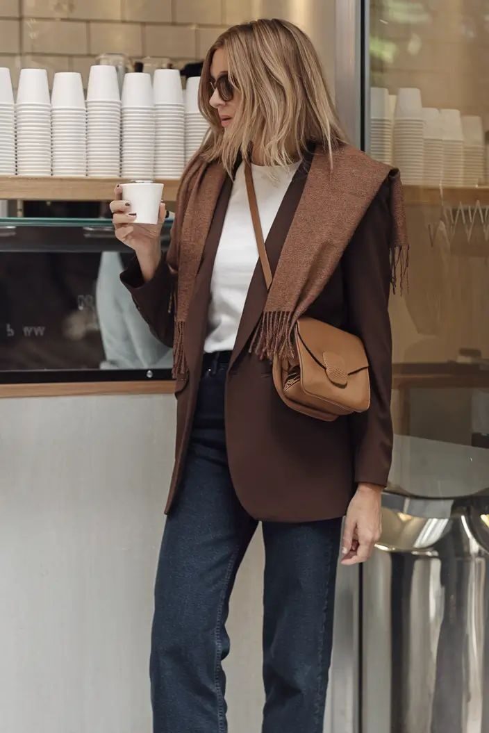 Trendy woman in sunglasses, blazer, and jeans enjoying coffee at a cafe. Casual chic style with sneakers and crossbody bag.