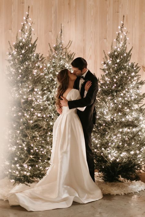 Bride and groom share a kiss in front of twinkling Christmas trees, capturing a magical winter wedding moment.