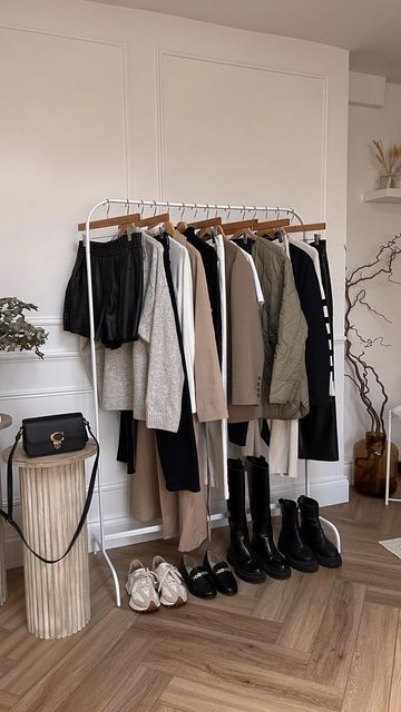 Clothing rack with neutral-toned outfits and black boots, accompanied by a small black handbag and beige shoes in a stylish room.