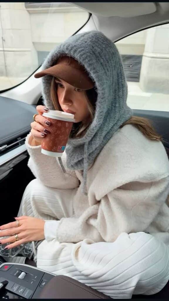 Person in cozy sweater and cap enjoys hot drink in car, winter fashion vibes.