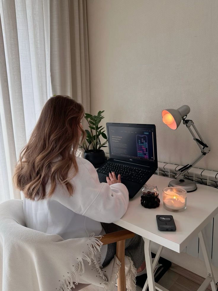 Cozy home workspace with woman coding on laptop, surrounded by plants, candles, and a lamp, promoting focus and relaxation.