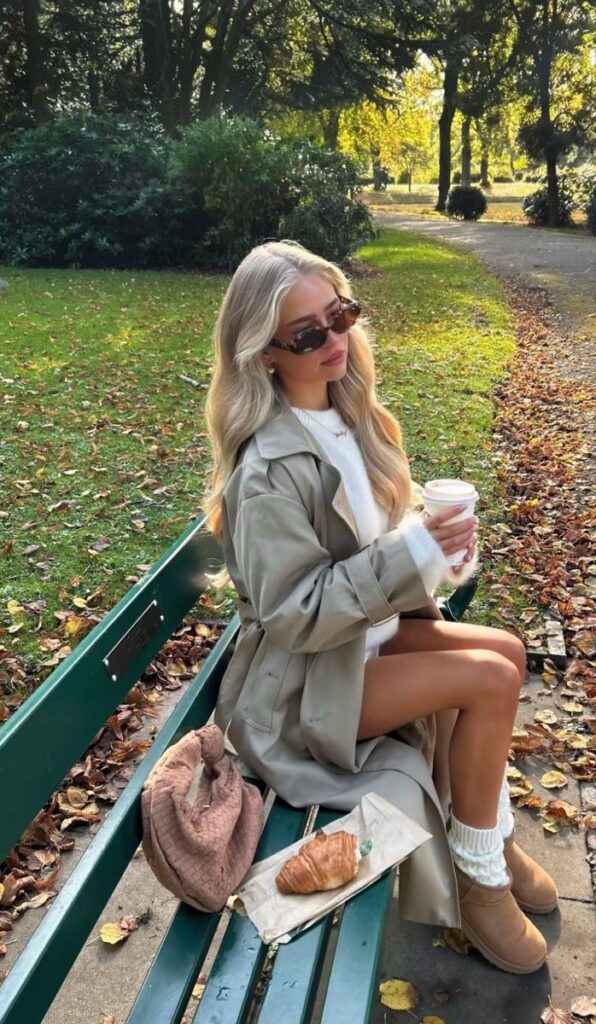 Woman in autumn coat sits on park bench, holding coffee with croissant nearby, enjoying a sunny fall day.