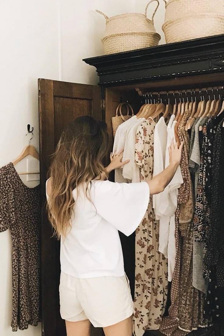 Woman browsing clothes in a wooden wardrobe, featuring floral and neutral dresses. Cozy and minimalist wardrobe setup.