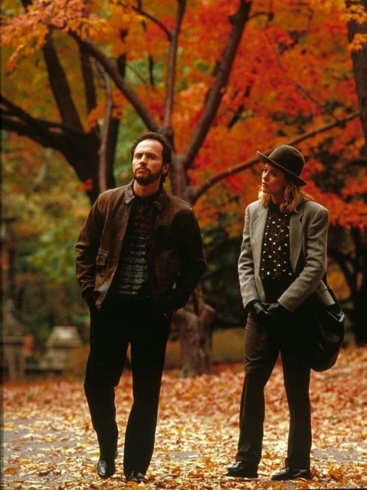 A man and woman walking in a park during autumn, surrounded by colorful foliage and fallen leaves.