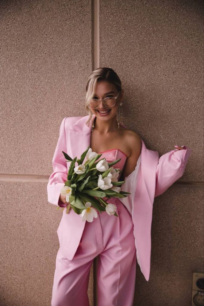 Woman in pink suit holding tulip bouquet, smiling against a textured wall. Fashionable spring style.