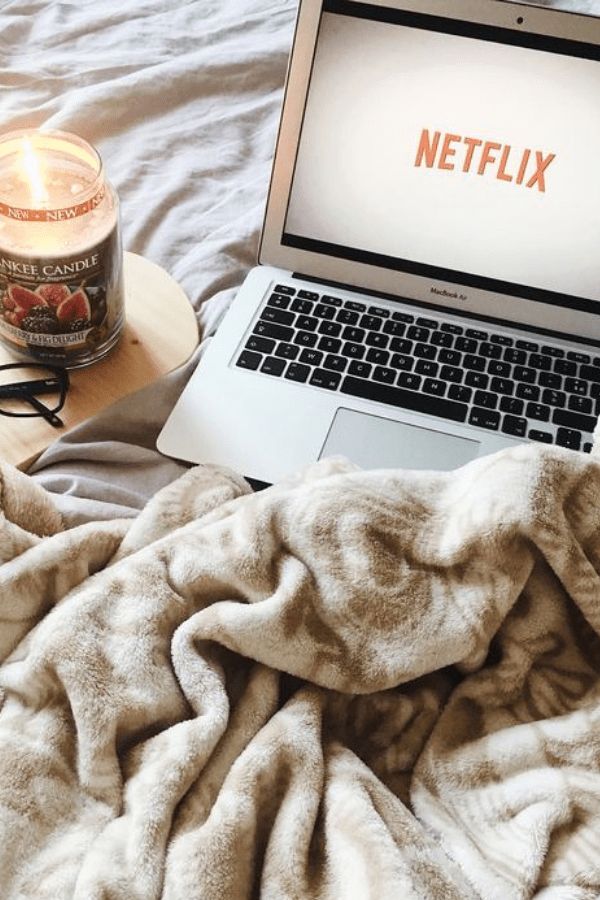 Cozy setup with Netflix on a laptop, a warm blanket, and a lit candle on a wooden table for a relaxing day in bed.