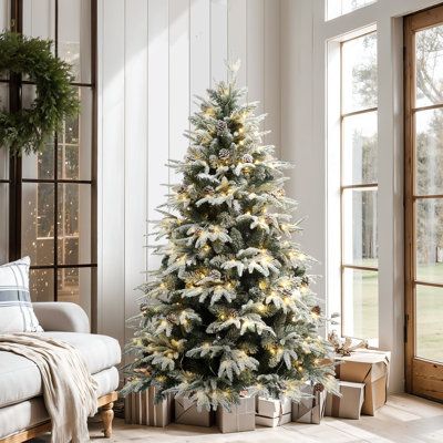 Decorated Christmas tree with gifts beneath in bright living room, featuring large windows and cozy seating area.
