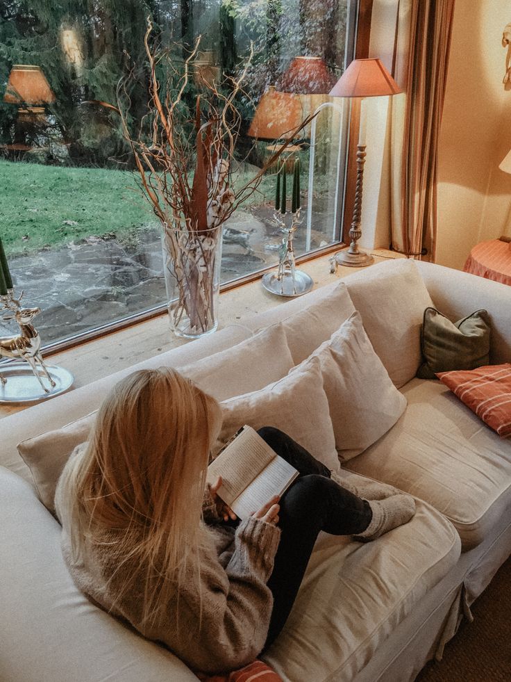 Cozy living room scene with a woman reading on a white sofa by a large window, surrounded by nature and warm decor.
