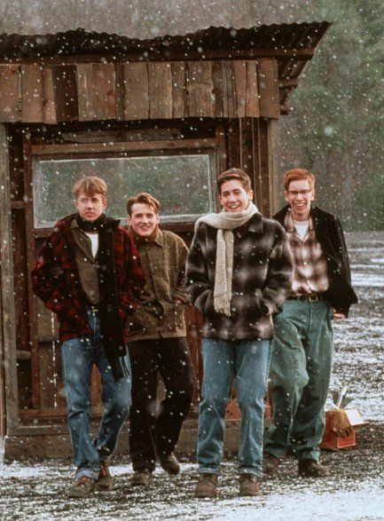 Four men smiling and walking in the snow, dressed in warm winter clothing, in front of a rustic wooden cabin.