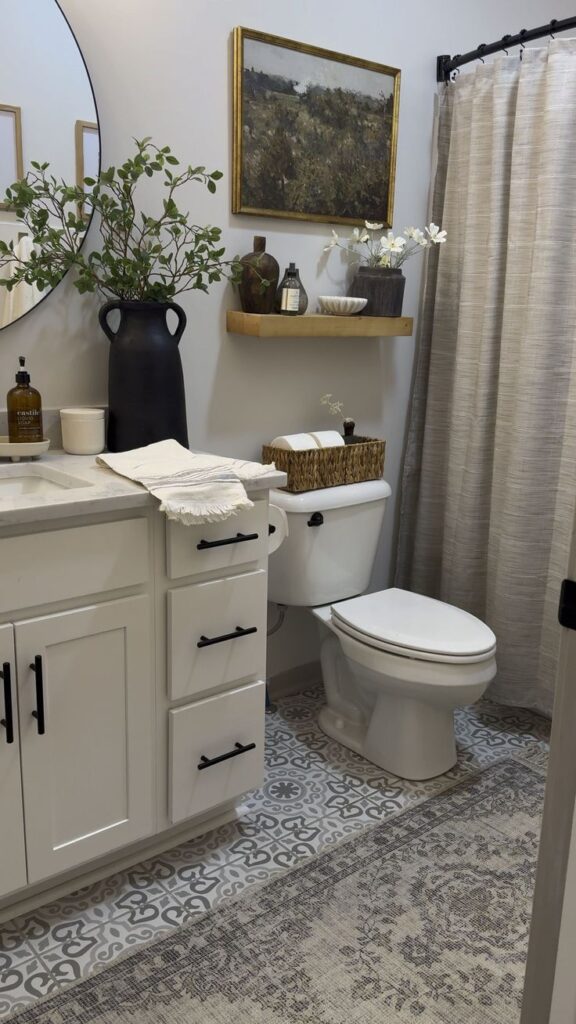 Modern bathroom with white vanity, round mirror, and rustic decoration. Features tile flooring, a woven basket, and greenery accents.