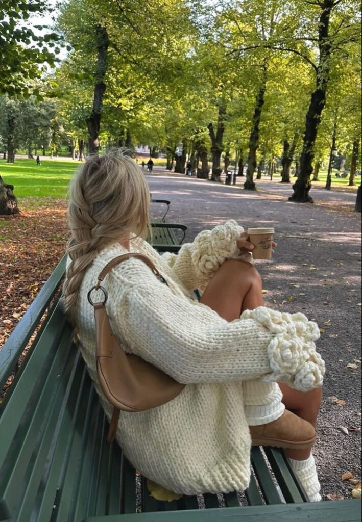 Woman in cozy knit sweater enjoys coffee on park bench in autumn. Relaxing in nature, serenity, fall fashion.