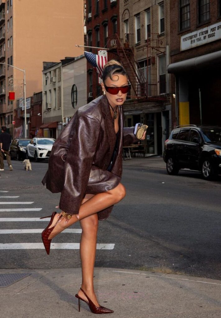 Fashionable woman in oversized leather jacket and heels poses stylishly on a city street corner. Urban fashion scene.