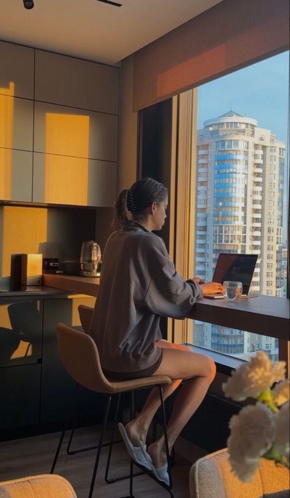 Woman working on laptop by window with city view, natural light filling modern kitchen space.