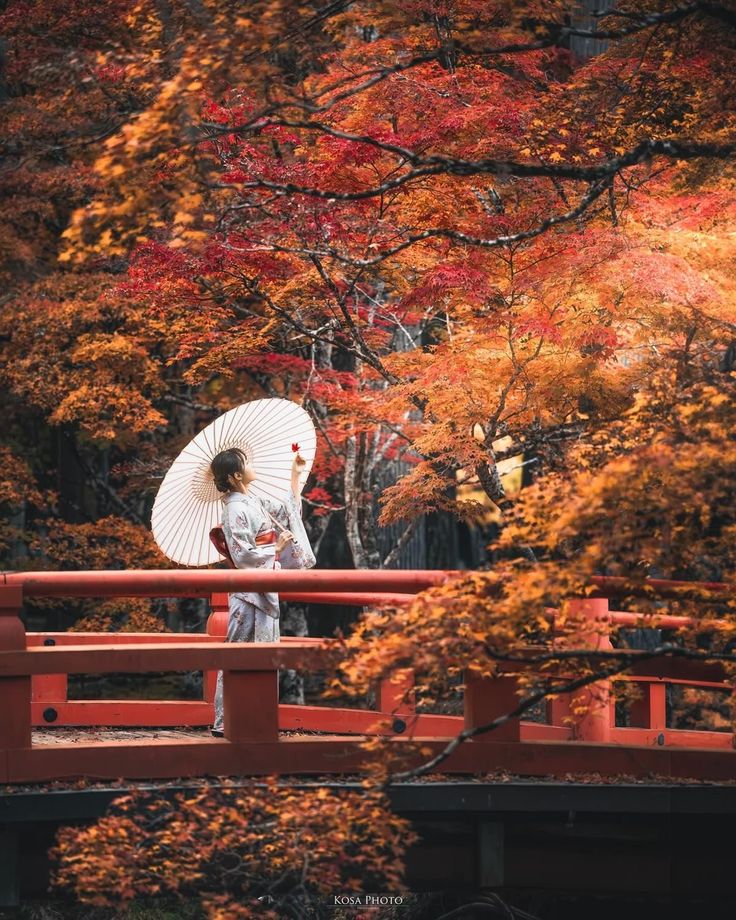 Person in traditional attire holding an umbrella, standing on a bridge surrounded by vibrant autumn leaves.