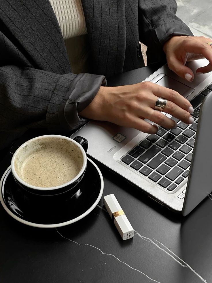 Woman typing on a laptop beside a coffee cup and lipstick, wearing a striped suit. Productivity and style in a café setting.