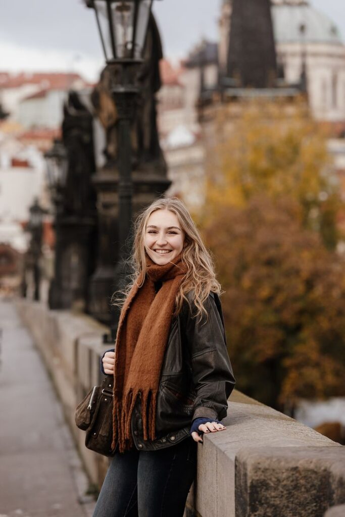 Smiling woman in a brown scarf and leather jacket on a scenic bridge, surrounded by autumn colors.