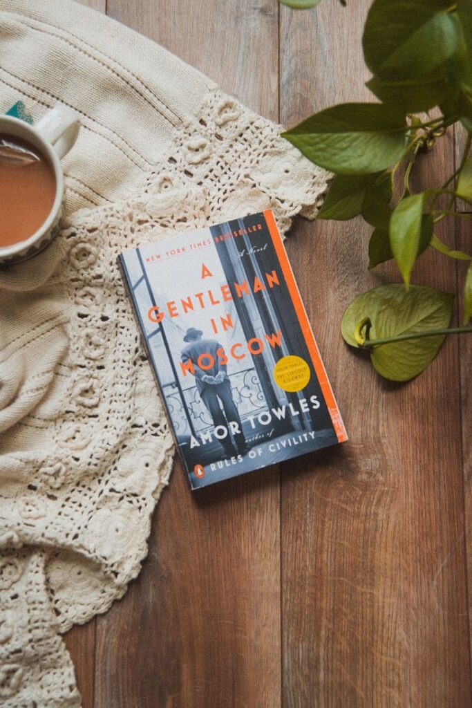 Book A Gentleman in Moscow by Amor Towles on a wooden table with a knit cloth, coffee, and plant. Cozy reading setting.
