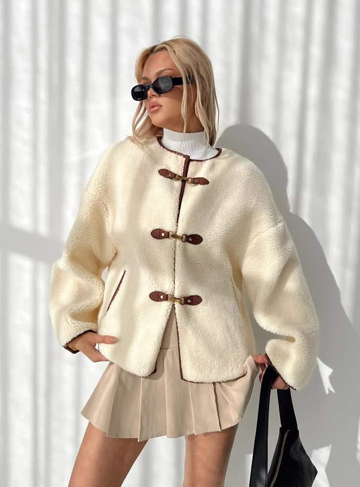 Stylish woman in a cozy shearling coat and pleated skirt, accessorized with sunglasses, posing confidently against a light background.