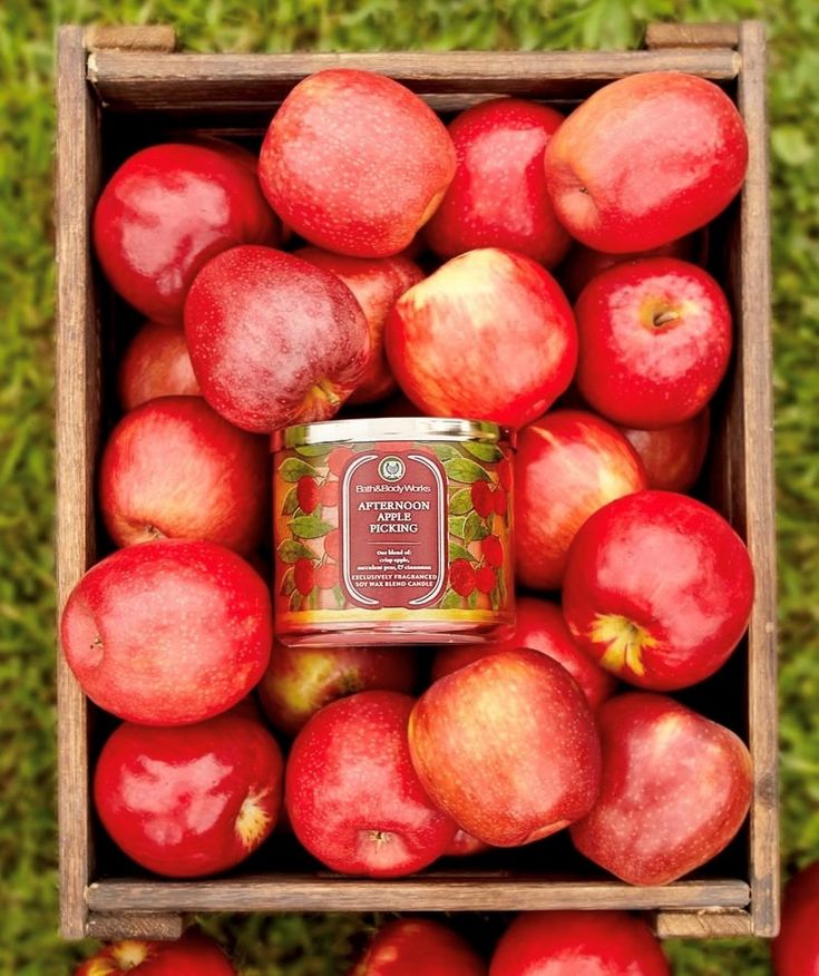 Box of fresh red apples with a candle labeled Afternoon Apple Picking centered, resting on a wooden crate, grassy background.