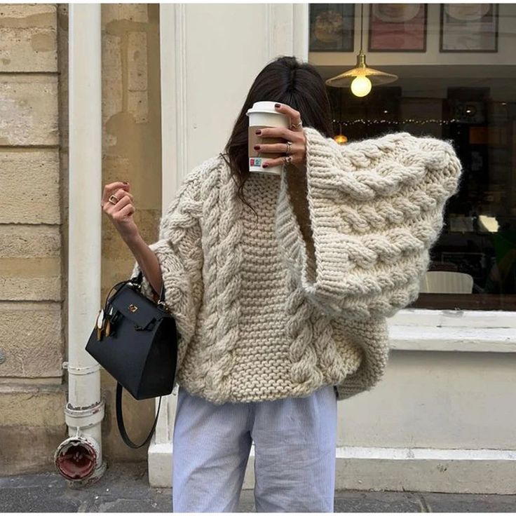 Woman in a chunky knit sweater holds coffee cup, stylishly accessorized with a black handbag, standing outside a café.