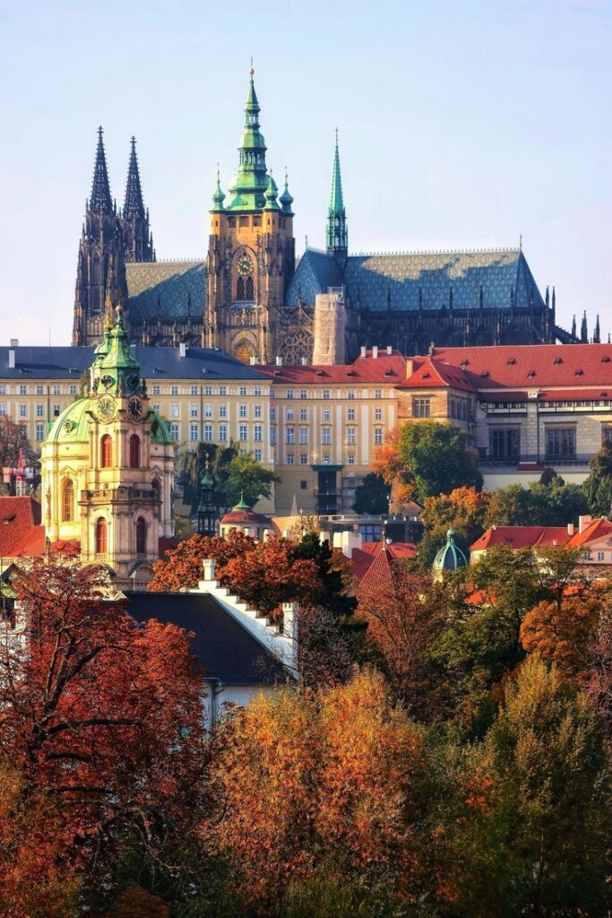 Stunning view of Prague Castle with colorful autumn trees, vibrant cityscape, and iconic architecture under blue skies.