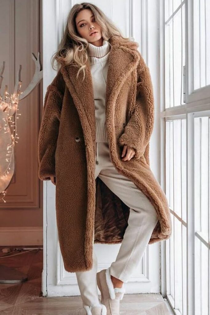 Woman in cozy brown winter coat and white sweater stands by window, showcasing trendy winter fashion indoors.