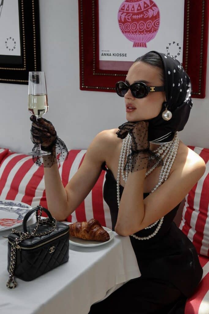 Elegant woman in vintage style sipping champagne, wearing pearls and sunglasses, at a chic cafe with a croissant and handbag.