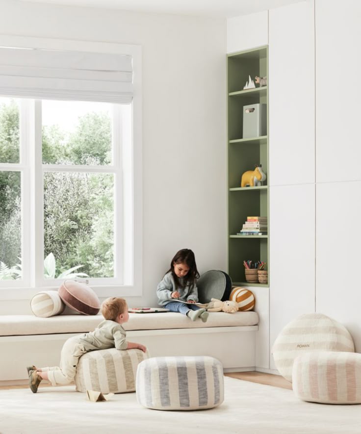 Children playing in bright, modern playroom with poufs, books, and natural light through large window. Cozy and inviting space.