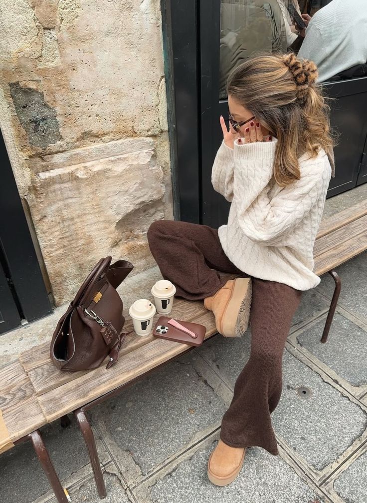 Woman in stylish winter outfit sitting outdoors with coffee cups and a brown bag on a bench, urban setting. Cozy fashion moment.