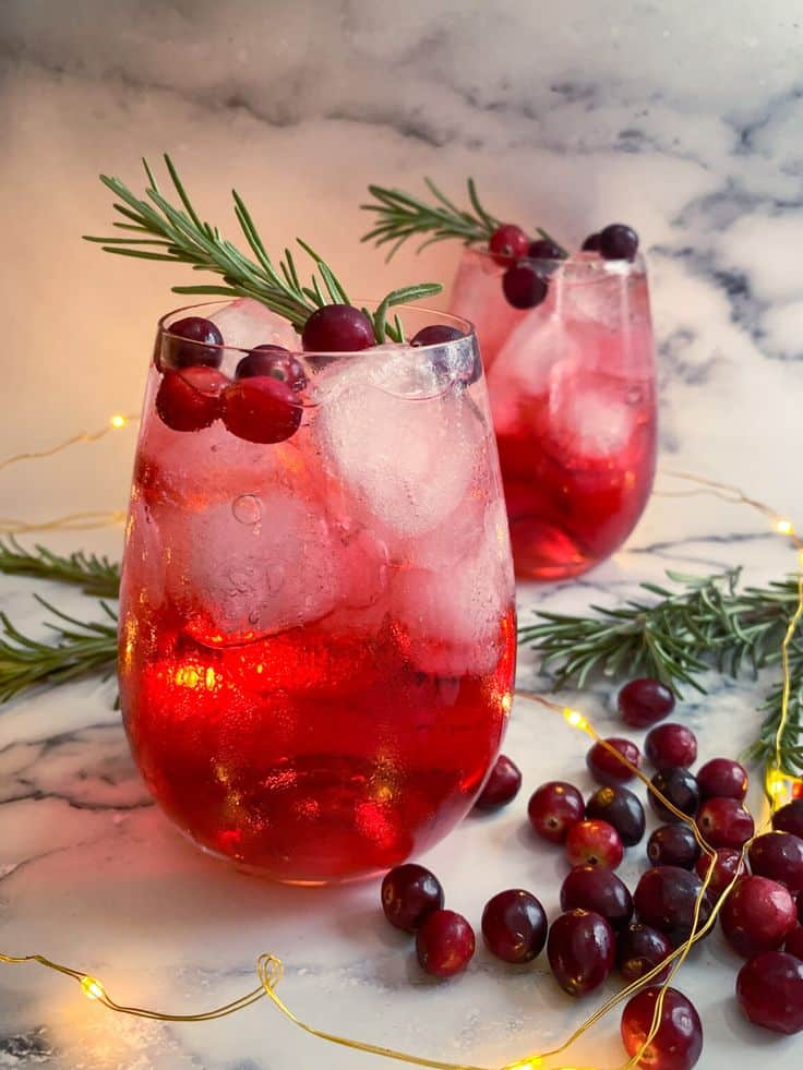 Festive cranberry cocktail with ice, garnished with rosemary, on a marble table with cranberries and lights. Perfect holiday drink.