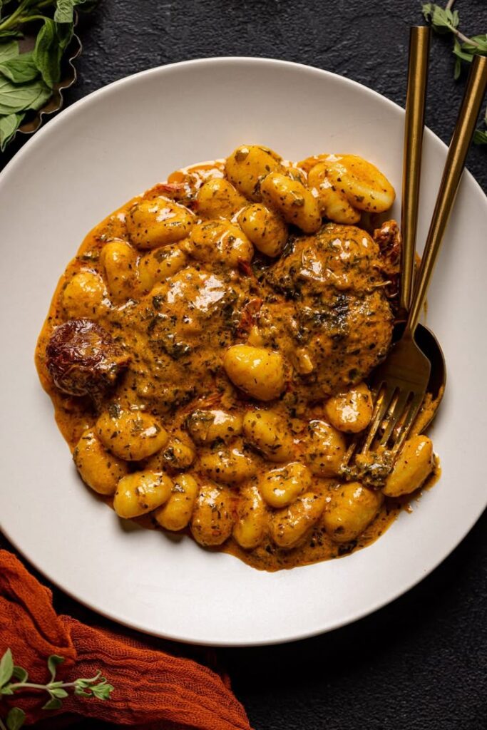 Creamy gnocchi with sun-dried tomatoes and herbs served in a white bowl, accompanied by a fork and spoon.