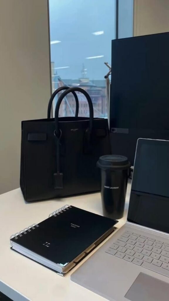 Modern workspace with a black handbag, spiral notebook, laptop, and coffee cup on a desk near a window view.