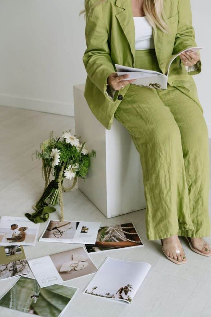 Person reading magazine in green suit, seated beside flowers and scattered open magazines on the floor, in a minimal setting.