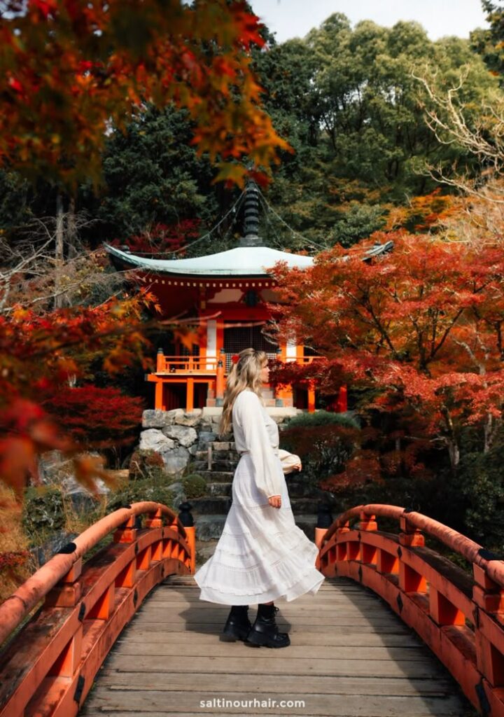 Woman in white dress standing on a red bridge with a traditional Japanese temple and vibrant autumn foliage in the background.