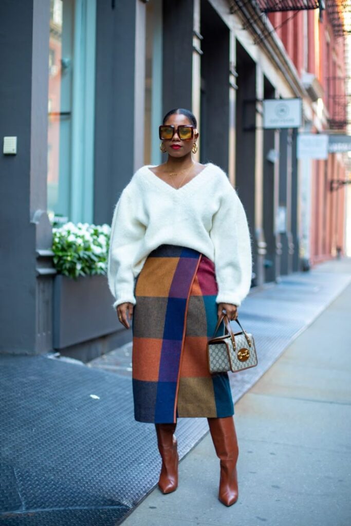 Stylish woman in a white sweater, colorful patchwork skirt, and brown boots, holding a designer bag on a city street.