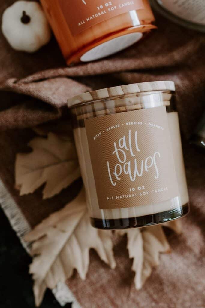 Natural soy candle labeled Fall Leaves with scents of musk, apples, berries, and oranges, placed on a rustic fabric backdrop.