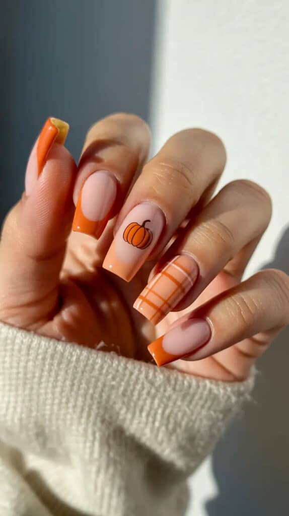 Fall-themed nails with orange tips, plaid design, and pumpkin accent on a cozy sweater background. Perfect autumn manicure inspiration.
