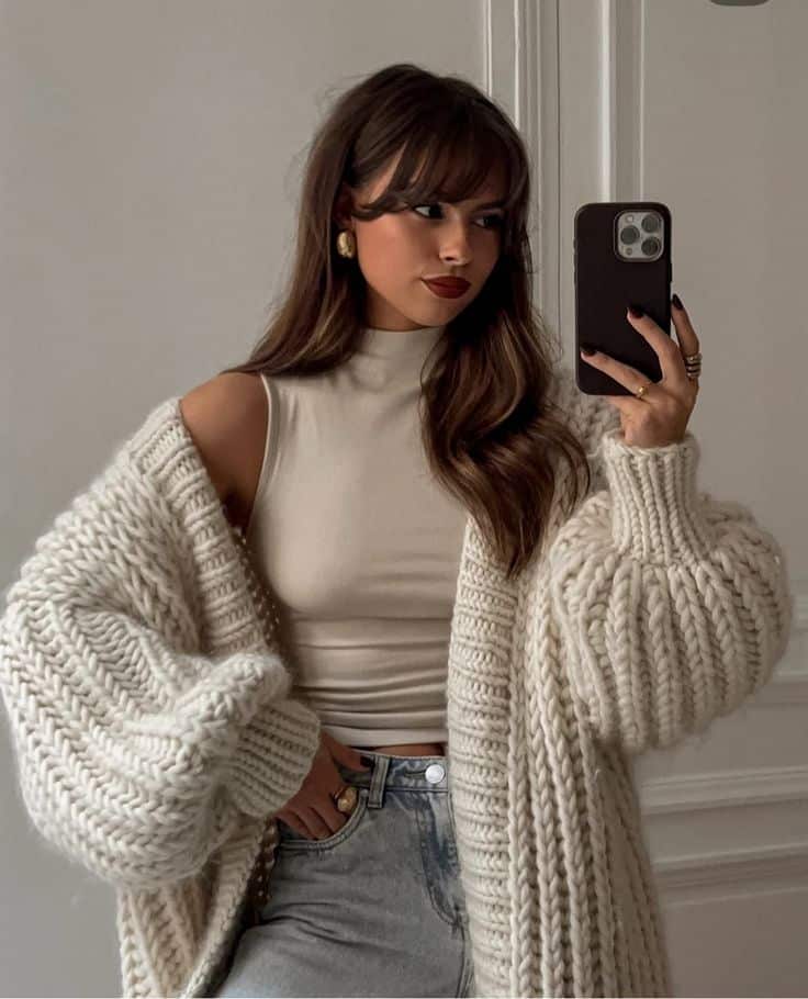 Woman in cozy knit cardigan takes a mirror selfie, wearing a sleeveless top and jeans. Elegant and stylish winter fashion.