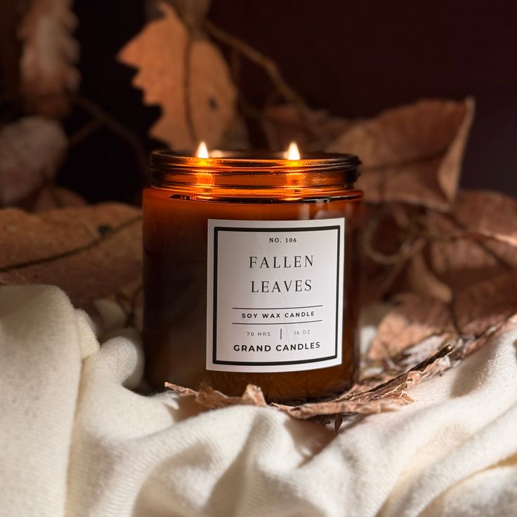 Amber jar candle labeled Fallen Leaves with a cozy autumn vibe, surrounded by dried leaves and soft fabric.
