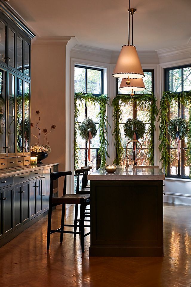 Modern kitchen with large windows, island counter, hanging pendant light, and festive greenery, providing a cozy and elegant atmosphere.