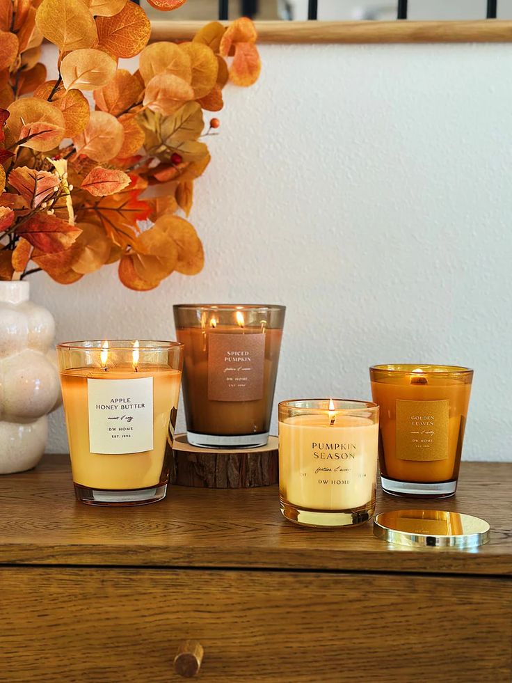 Autumn-themed candles on a wooden table with orange leaves in a vase, creating a cozy and warm seasonal atmosphere.