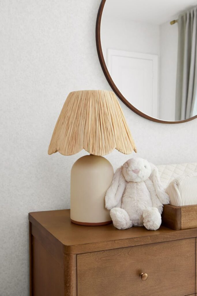Natural wood dresser with a round mirror, a soft toy bunny, a beige lamp with woven shade; cozy, minimalist decor.