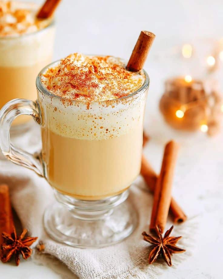 Creamy pumpkin spice latte with whipped cream, cinnamon stick, and star anise in a clear glass mug on a cozy table setting.