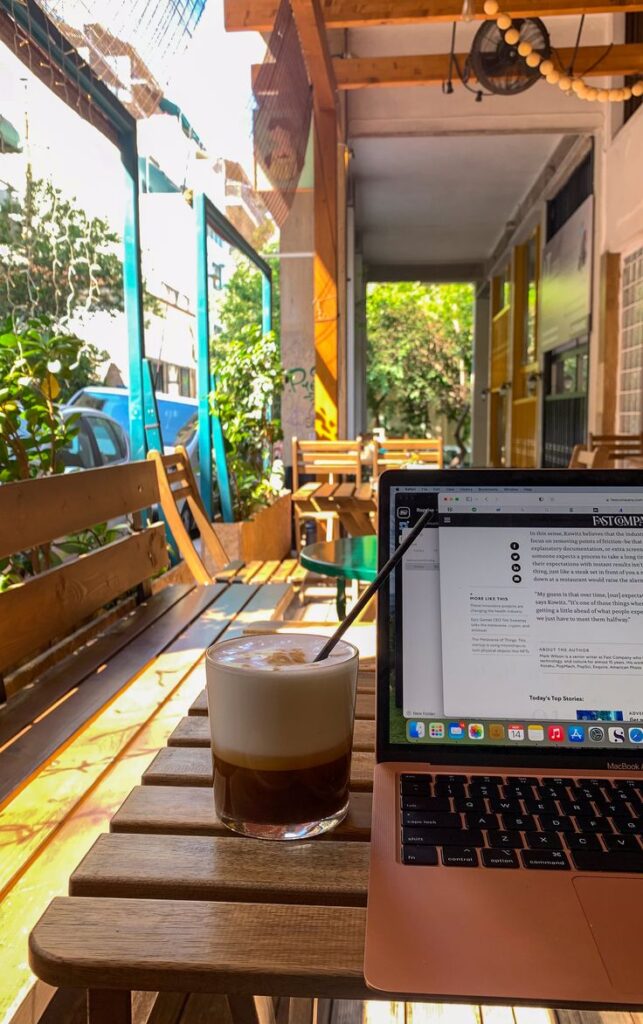 A cozy cafe setup with a laptop and iced coffee on a wooden table, bathed in sunlight through a window. Perfect for remote work.