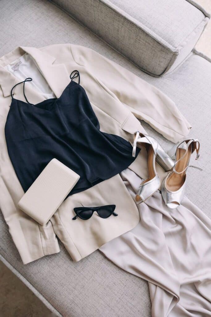 Elegant outfit with a beige blazer, navy camisole, clutch, sunglasses, and silver heels on a sofa. Perfect for sophisticated events.
