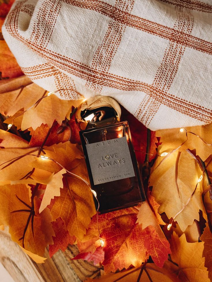 Perfume bottle with Love Always label on autumn leaves, cozy plaid fabric, and warm fairy lights for a fall-themed ambiance.