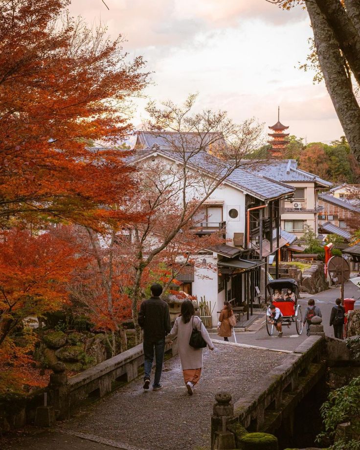 Couple walking in picturesque Japanese village with vibrant autumn leaves, traditional buildings, and a distant pagoda.