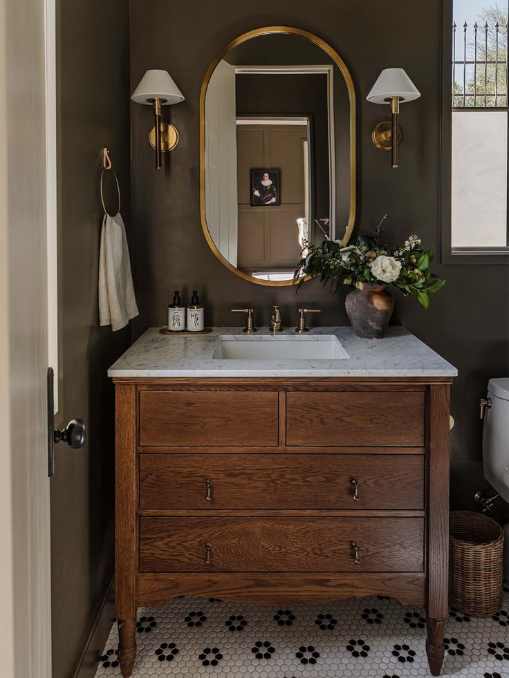 Elegant bathroom with vintage wood vanity, marble countertop, brass fixtures, oval mirror, and potted greenery.