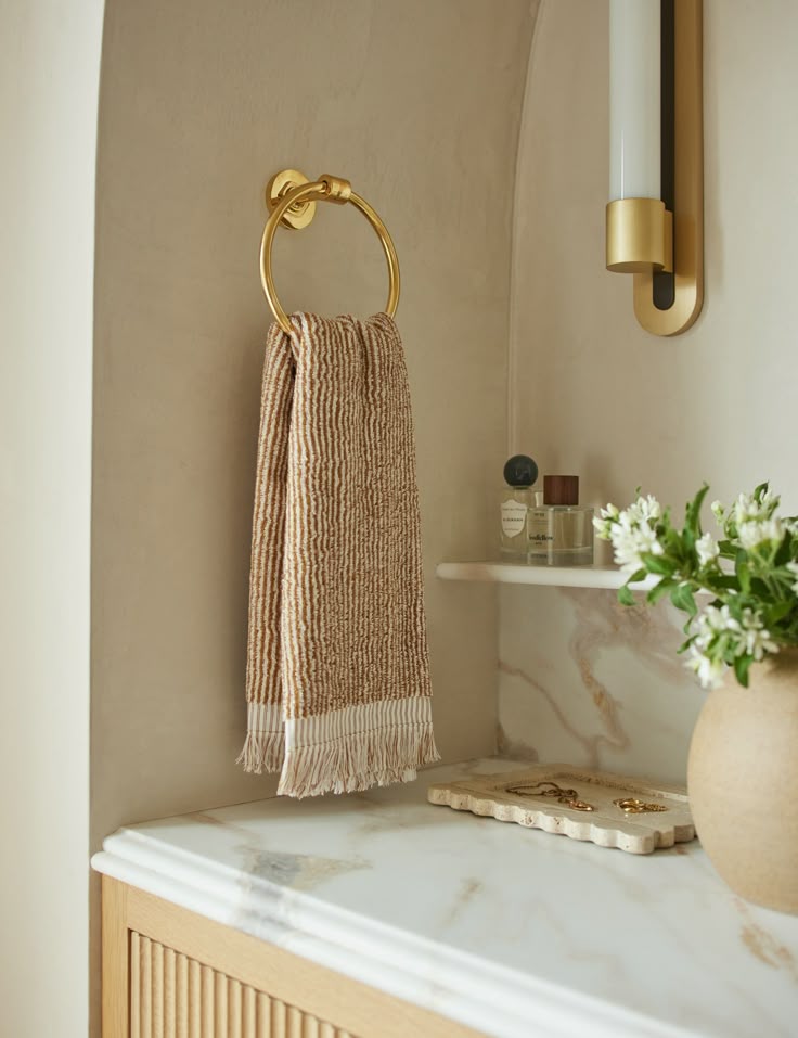 Elegant bathroom with gold towel ring, striped towel, and marble countertop. Decor includes a vase and toiletries on a shelf.